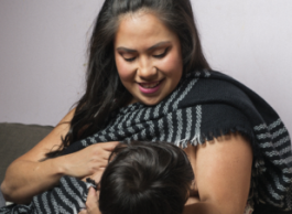 A woman with long black hair wearing a grey and black shawl breastfeeds a baby with black hair