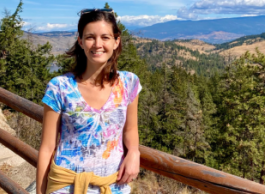 Smiling woman with long, brown hair, wearing a colourful, short-sleeved v-neck top, leaning on a wooden fence, overlooking a canyon with evergreen trees, mountains and clouds in the background