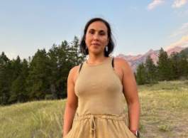 Woman in tan tank top standing on grass with mountains in background