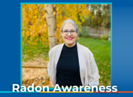 A smiling woman with curly grey hair, glasses, and red lipstick and wearing a black t-shirt and beige sweater stands with her hands in her pockets in front of a tree with yellow leaves.