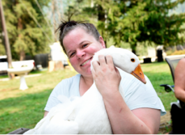 A woman holds a goose while sitting outdoors in a grassy area.