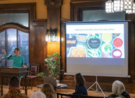 A woman in a green shirt gestures at a podium to the left of a screen showing pictures of food