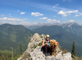 A woman with a yellow bandana and teal tank top sits with an orange dog on a rocky mountain with mountains and blue sky in the background