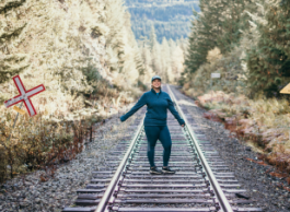 A woman in a blue track suit and hat stands on train tracks in the middle of a forest.