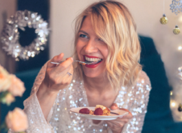 Blond woman in a silver sparkly outfit smiles and takes a bite out of a holiday dessert