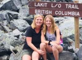 Blonde women with long hair smile and sit together on grey rocks with trail sign behind them