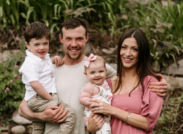 A man in a grey shirt and a woman with long dark hair in a pink dress stand together holding a baby and young child in a forest