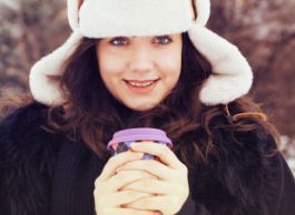 : a smiling brunette with a winter hat holds a coffee in her hand