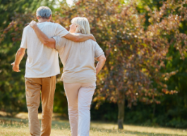 A woman and man both with grey hair wearing casual clothes walk arm in arm in a park on a sunny day