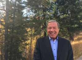 A smiling man with grey hair, wearing a suit jacket and button-up blue shirt, poses for a picture outside with trees and shrubs in the background. 