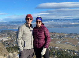 Two people wearing winter clothing stand at the top of a mountain with a city and blue sky in the background