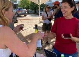 A blonde woman in a white tank top pours non-alcoholic wine into a cup for a smiling woman in a red t-shirt at an outdoor event