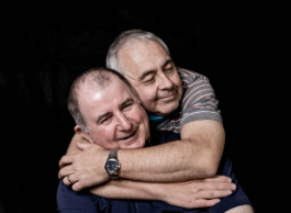 A man with greying hair wearing a striped grey shirt embraces a smiling man with black thinning hair