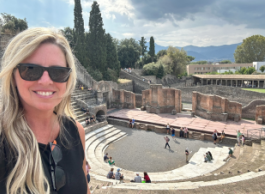 Smiling woman with long blonde hair and sunglasses at roman theatre with people, trees and mountains in background 