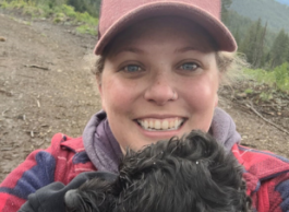 A blonde woman wearing a ball-cap smiles at the camera with her black dog