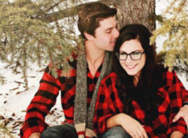 A woman and a man in red plaid shirts pose under a fir tree