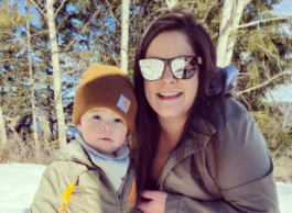 A woman with long brown hair and mirrored sunglasses kneels next to a young boy in a yellow toque on the edge of a snowy forest