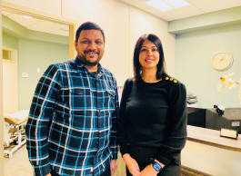 A smiling man with black hair and beard in a blue plaid shirt stands next to a smiling woman with long black hair wearing a black top