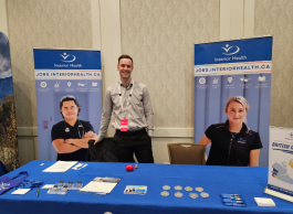 A smiling man with short brown hair wearing a light grey dress shirt and red lanyard stands behind a table with a blue tablecloth between two large posters of health care workers at a job fair