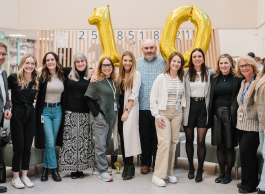 A group of 12 people wearing business attire stand together arm in arm in an office with gold mylar balloons that say 10 behind them