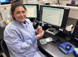 Smiling woman with brown hair and blue lab coat sitting at computer desk in front of monitor holding test swab