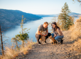 A young couple in casual clothes kneels on a gravel path with their small caramel coloured dogs with a lake, mountains and trees in the background on a sunny day