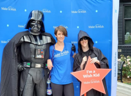 A woman in a blue t-shirt with short brown hair stands arm in arm with a young man wearing a black cloak and holding a red cardboard star and a person in a Darth Vader costume with black armour, helmet and cape