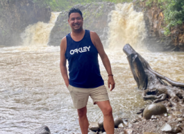 A smiling man with dark hair wearing a blue tank top that says Oakley stands in sandals in front of two waterfalls on a rocky beach.