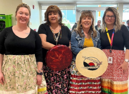 Four smiling women stand next to each other all wearing bright ribbon skirts, and black shirts or jean jackets. Two are holding Indigenous drums.
