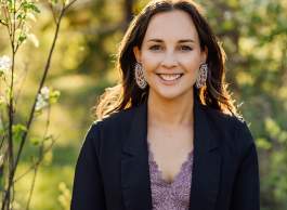 Indigenous woman, shoulder length brown hair wearing beaded Indigenous-designed earrings, smiling outdoors in front of budding shrubbery known as the Saskatoon. 