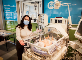 A woman with long dark hair and cream coloured sweater and wearing a face mask stands next to a baby bassinette inside a neonatal intensive care unit at a hospital