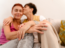 Two older women sitting on a couch in each other's arms, one with short red hair and a pink tanktop, and one with short dark hair with a white shirt, beige pants and big silver bracelet. She's kissing her partner on the forehead