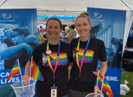 Two women with their hair pulled back in ponytails smile at the camera. They wear Pride rainbow t-shirts, Interior Health rainbow lanyards, and stand in front of Interior Health banners. There are rainbow flags in the foreground. 