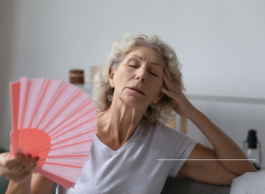 An older woman with long grey hair with her head in her hand and eyes closed fans herself with a red fan