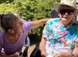 An older woman in a pink tshirt puts her arm around an elderly man wearing a bright multi-coloured tshirt, sunglasses and hat