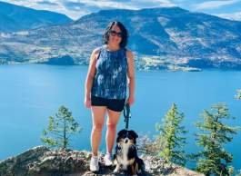 Woman in blue tank top and black shorts standing on a mountain with her dog and a lake in the background