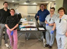 Five people standing around a table, two on left, three on right, with a stack of small boxes and information posters for medical gloves. Three of the five are either wearing or holding blue medical gloves.