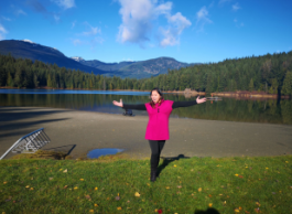 Woman in pink shirt stands with arms open in front of a beautiful mountain and lake scenery 