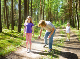 A middle aged person sprays insect repellent on a child's leg as they stand on a forest path. Another child stands behind them and watches.