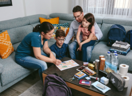 Two adults and two children sit on a couch around a table reviewing a document. The table has a first aid kit on it and emergency supplies such as bottled water, toilet paper and food items.