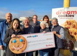 A group of seven smiling people wearing fall clothing stand in the sun holding a cheque with a large cookie on it in front of a sign that says new dream cookies