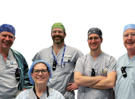 Five smiling health-care workers in medical clothing, four standing and one kneeling in front, all wearing surgical caps and ID badges.