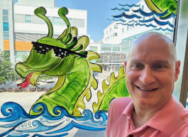 A man wearing a business casual shirt poses by a window that has a mural of a sea dragon wearing sunglasses.