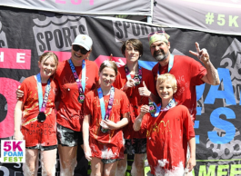 Two adults and four preteen children in red shirts with medals around their neck pose in front of a black sign with pink and blue lettering.