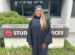 A woman wearing graduation attire poses in front of a college on a bright day. 