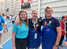 Photo of blonde-haired woman smiling for the camera with her son and daughter. She is between them, wearing a black T-shirt and blue jeans with a dark blue apron and a rainbow lanyard. On her left her daughter with blonde hair is smiling and wearing a teal blue T-shirt and black jeans. To the left, her blonde-haired son with black rimmed glasses is wearing a dark blue T-shirt and a dark blue apron.