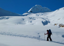 A person with a backpack skis across a snow-covered landscape toward a distant mountain peak under a clear blue sky.