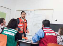 Five adults wearing vests labeled with various roles such as 'Operations' and 'Safety' are participating in a meeting led by a person standing in front of a whiteboard. The whiteboard includes a list of bullet points under the heading "Plan &amp; Budget".