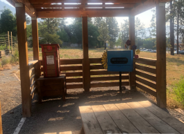 An outdoor covered wooden shelter with two informational signs displayed prominently. One red informational sign is affixed to the left, while a blue sign saying "Seed Exchange" is mounted on the front rail. The shelter has a simple, wooden bench beneath it. The setting is a sunny day with trees and a wire fence visible in the background.