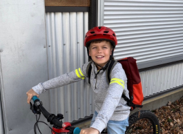 A child wearing a red helmet and a backpack smiles while sitting on a bicycle near a metal wall.